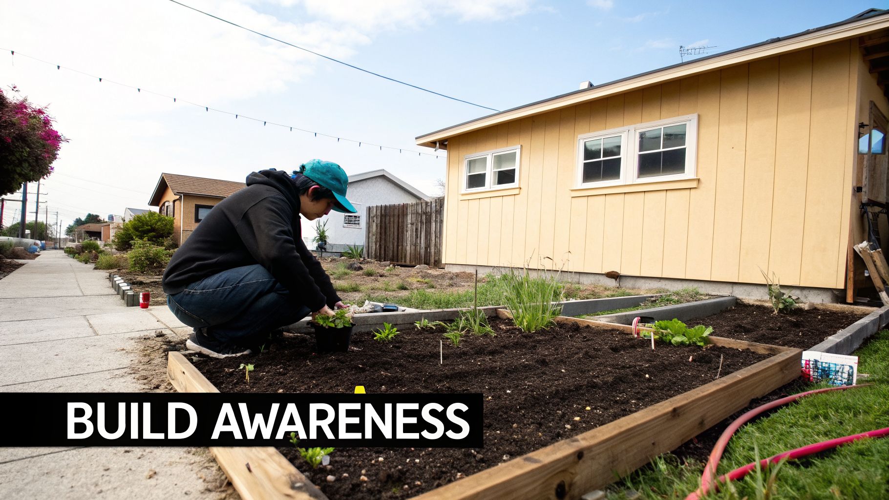 A person in a black hoodie and blue hat plants seedlings in a raised garden bed next to a yellow house.